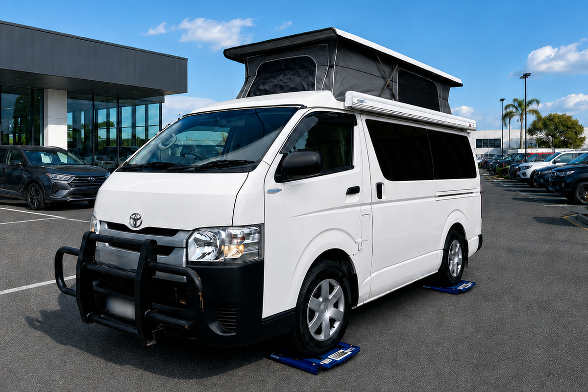 A white Toyota Hiace van with a black pop-up roof tent and a black front bull bar, parked in an outdoor dealership lot under a blue sky.