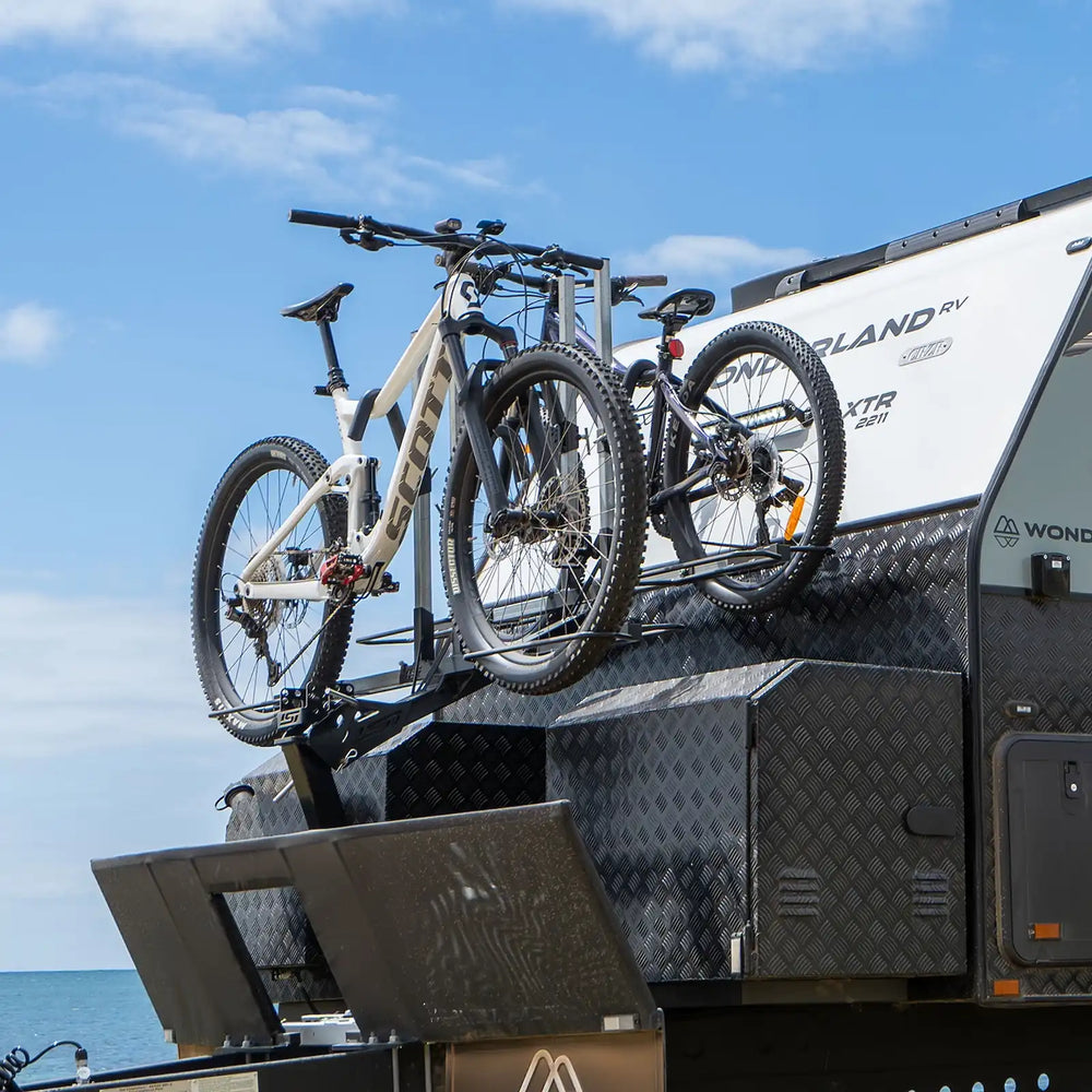 Two mountain bikes mounted on a roof bike rack of a white and black awning trailer, with the ocean and blue sky in the background.