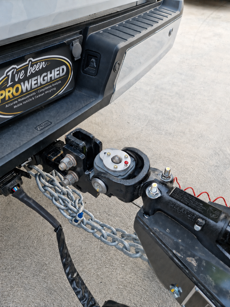Close-up of a black vehicle hitch and chain connection at the rear of a truck, with a visible 'I've been PROWEIGHED' badge on the bumper and concrete ground in the background.