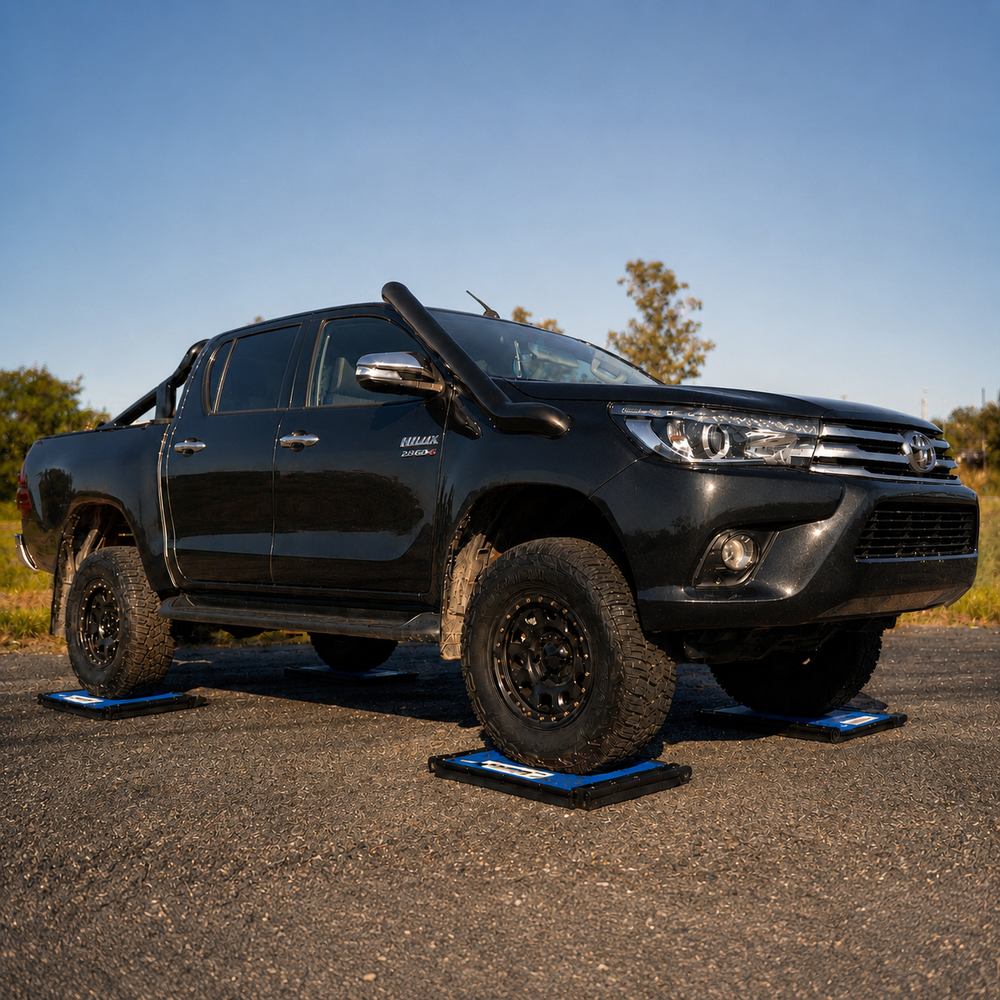 A black Toyota Hilar pickup truck parked outdoors on asphalt, with blue and black wheel chocks visible under each tire. The truck has off-road tires, a snorkel, and side mirrors, with trees and blue sky in the background.