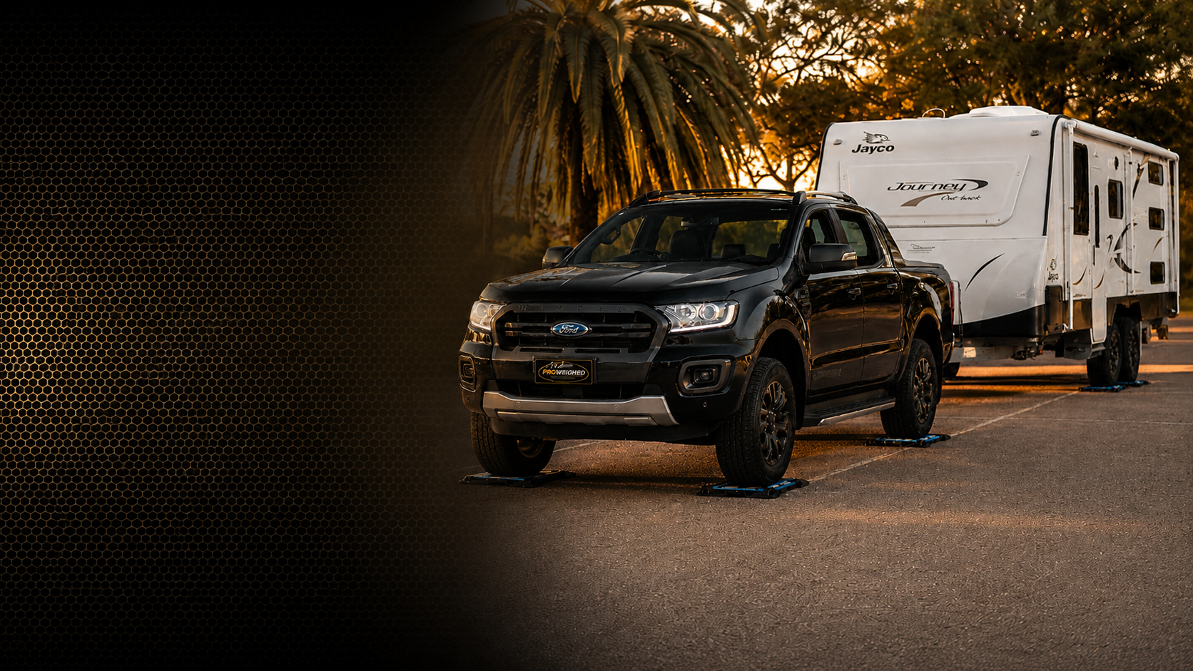 A black Ford Ranger pickup truck parked outdoors on a paved road, towing a large white travel trailer. The scene is lit by warm golden-hour sunlight with palm trees and trees in the background.