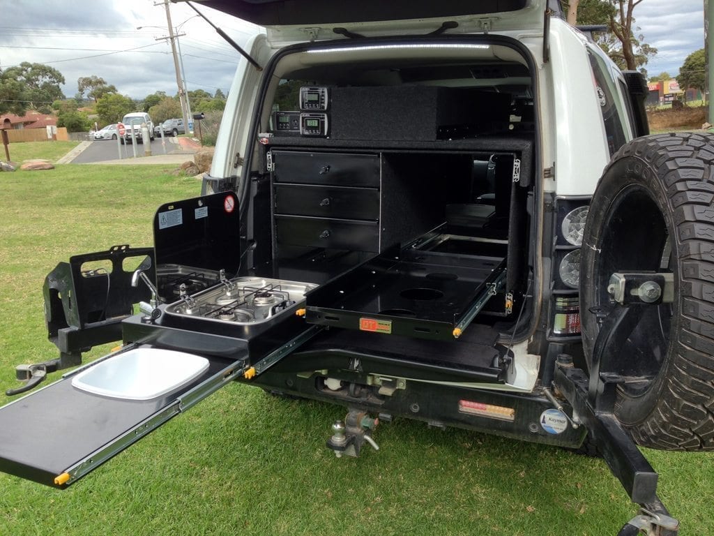 A black outdoor vehicle kitchen and storage system installed in the rear cargo area of a white off-road vehicle, shown with drawers, a slide-out work surface, a built-in stove, and a fold-down tray on grass outdoors.