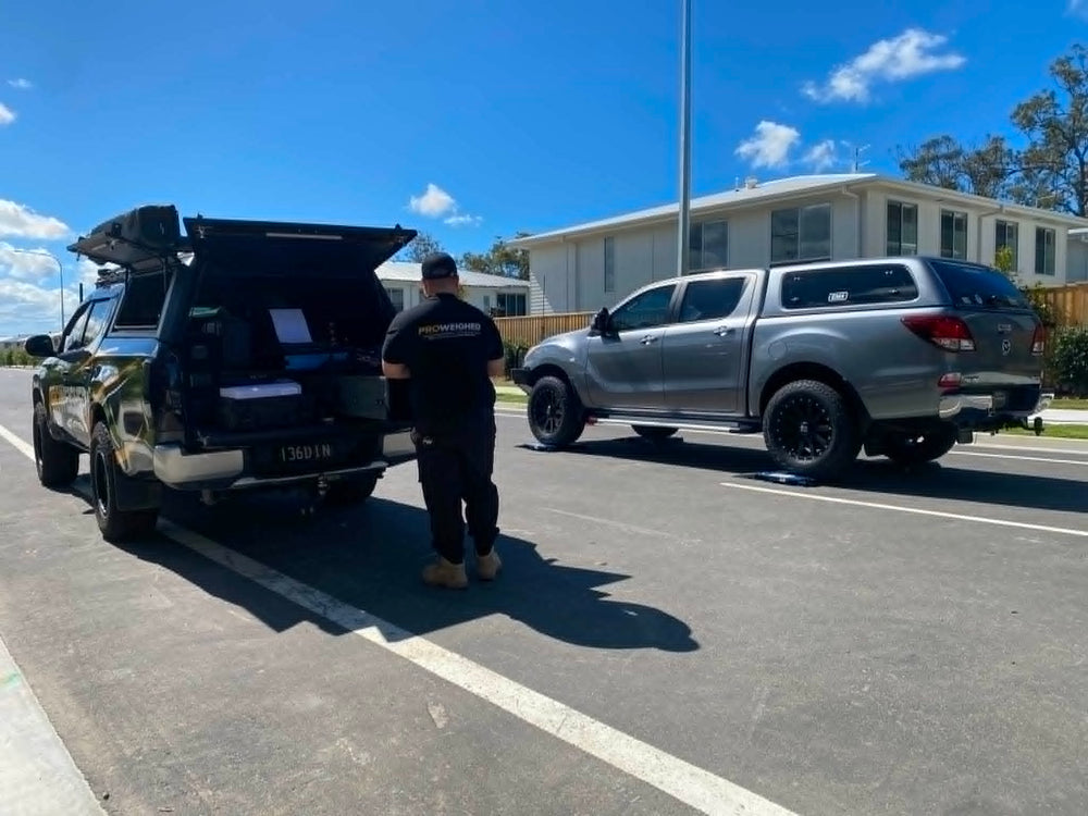 A man in a black t-shirt and dark pants stands between two vehicles parked on a street. On the left is a black pickup truck with its rear hatch open, and on the right is a silver pickup truck.