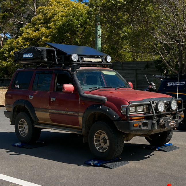 A red lifted SUV and a black branded pickup truck parked outdoors on asphalt, with orange cones in the foreground and trees and a building in the background.