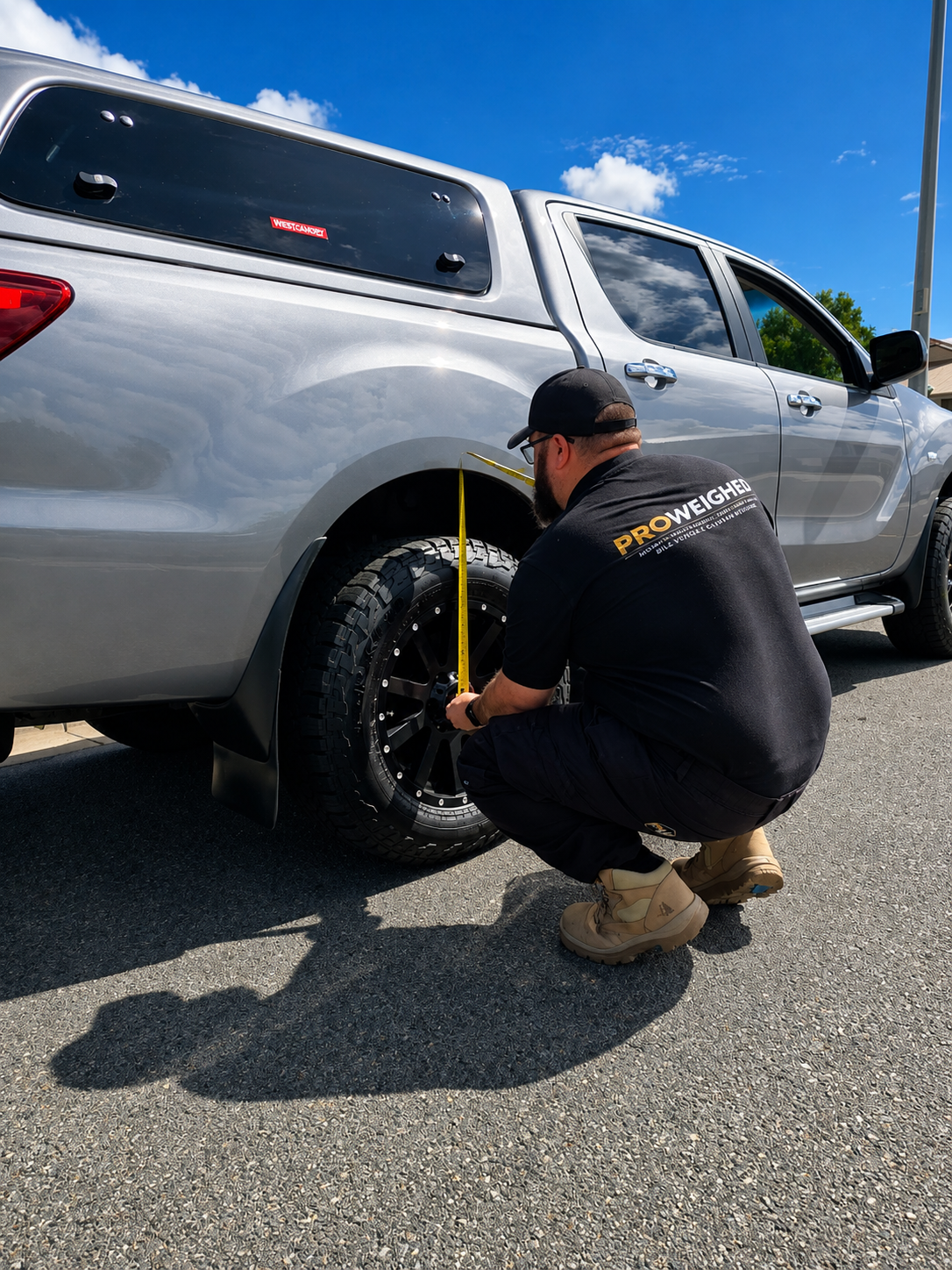 A man in a black branded t-shirt and cap crouches beside a silver pickup truck, measuring the tire width with a yellow tape measure outdoors on asphalt under a blue sky.