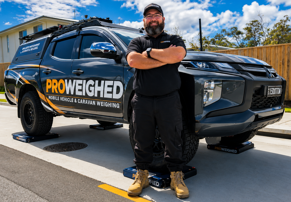 A man stands with arms crossed in front of a dark gray pickup truck with 'PROWEIGHED' branding, parked on a street with vehicle scales visible under the truck. The scene is set under a bright blue sky with white clouds.