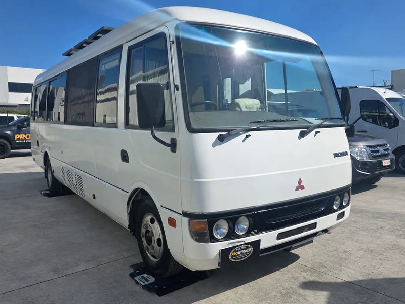 A white Mitsubishi Minivan bus parked on a concrete lot under a clear blue sky, with other vehicles visible in the background.