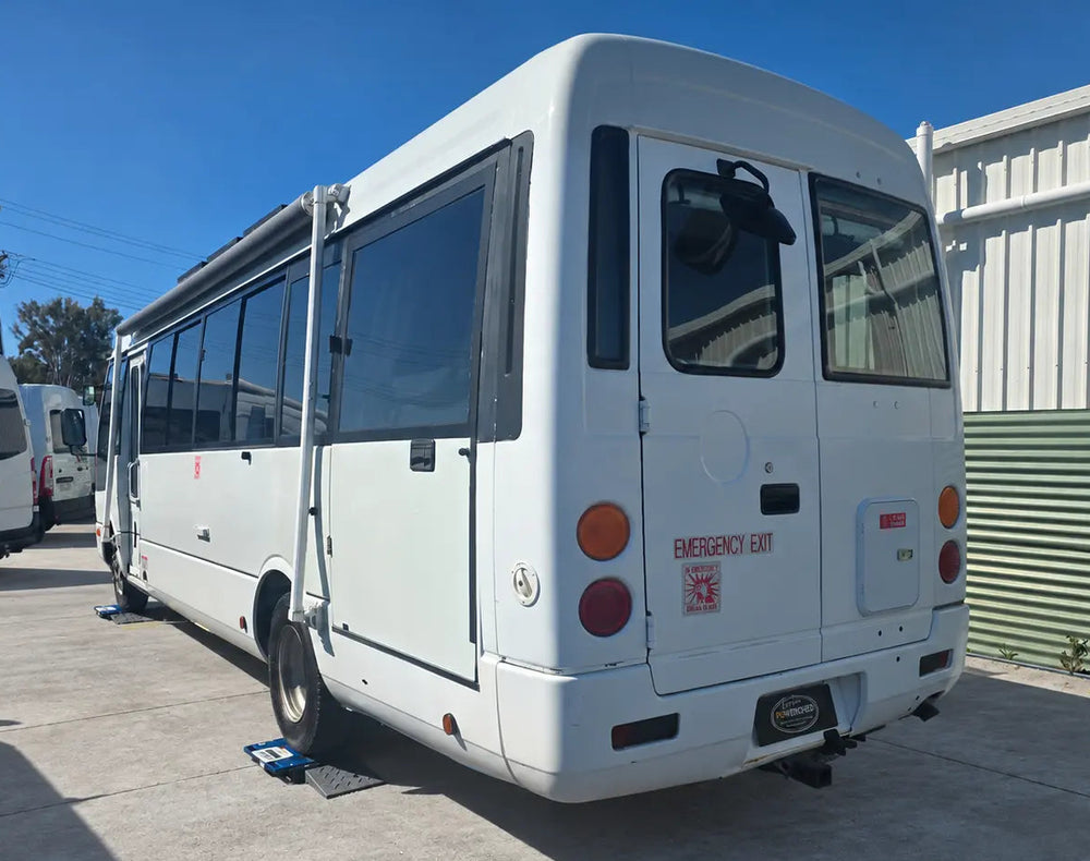 A white emergency response shuttle bus parked outdoors on a concrete lot next to a green corrugated metal building under a clear blue sky.