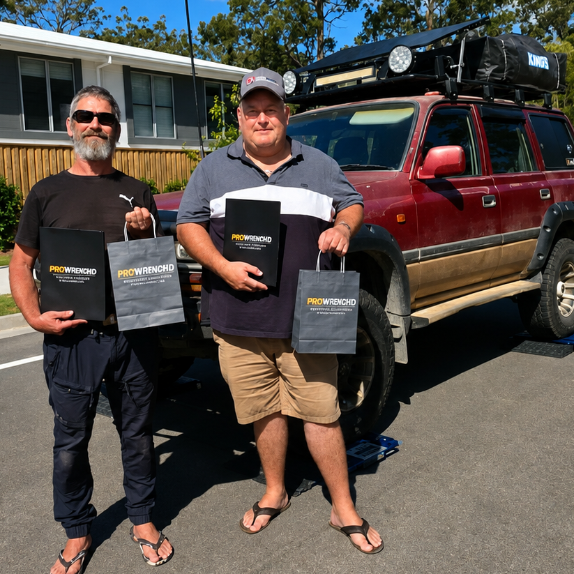Two men standing outdoors in front of a red SUV, each holding a black branded shopping bag. A house, trees, and blue sky are visible in the background.