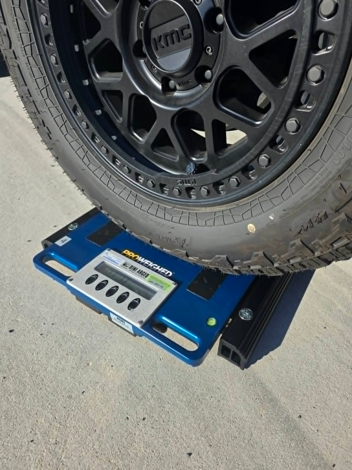A blue Dini Argeo vehicle scale positioned under a black off-road tire on an asphalt surface.