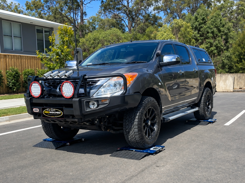 A black off-road pickup truck with a front bull bar and auxiliary lights is shown in an outdoor paved lot with orange cones, trees, and a house in the background.